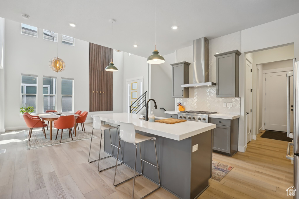 Kitchen featuring gray cabinetry, wall chimney range hood, light countertops, light wood-style flooring, and a kitchen island with sink