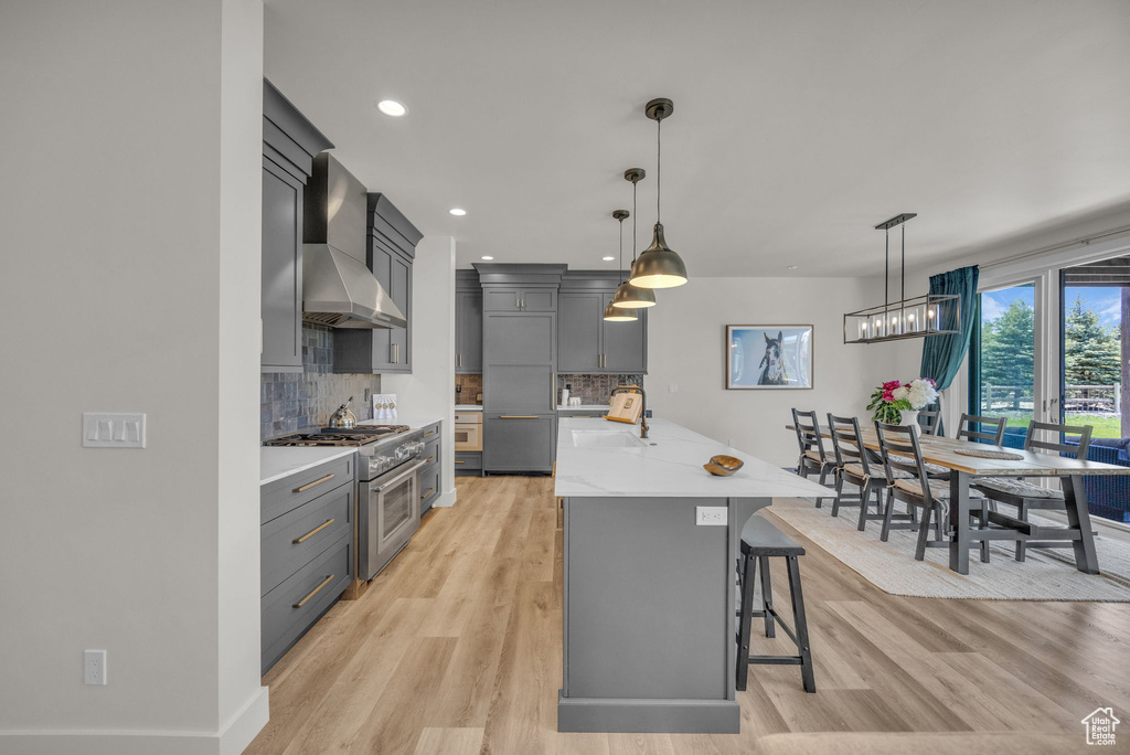 Kitchen with high end stove, wall chimney exhaust hood, light wood-style floors, an island with sink, and recessed lighting
