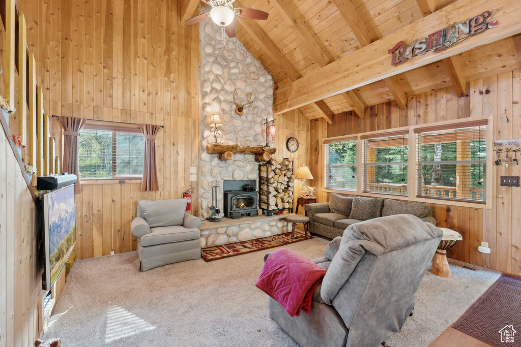Carpeted living room featuring healthy amount of natural light, wood ceiling with exposed beams, a wood stove, wooden walls, and a ceiling fan