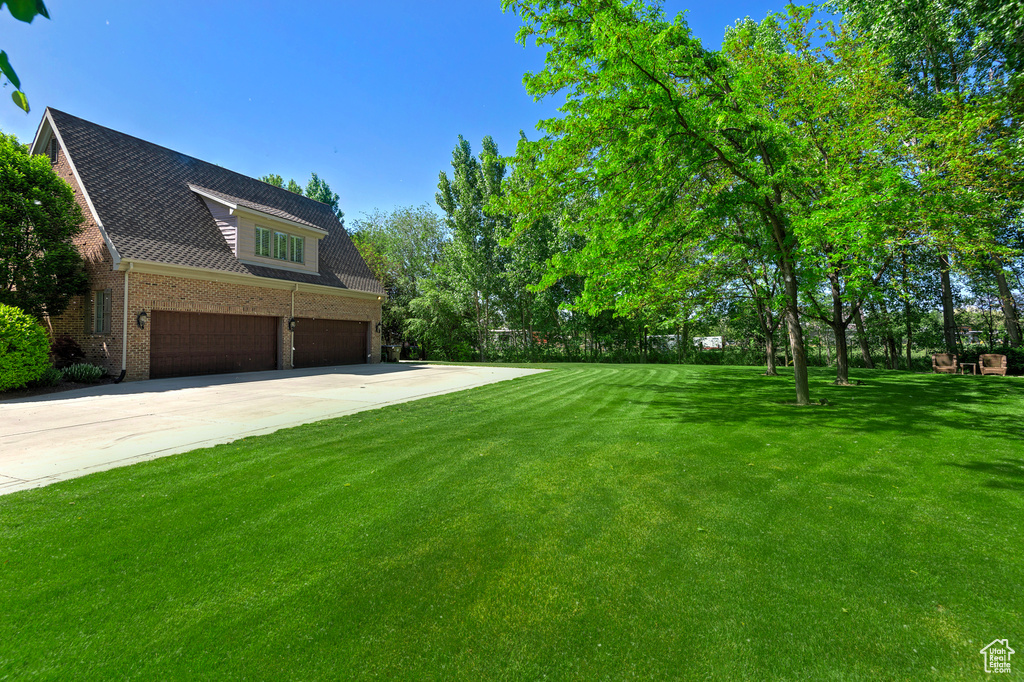 View of green lawn with a garage, concrete driveway, and view of wooded area