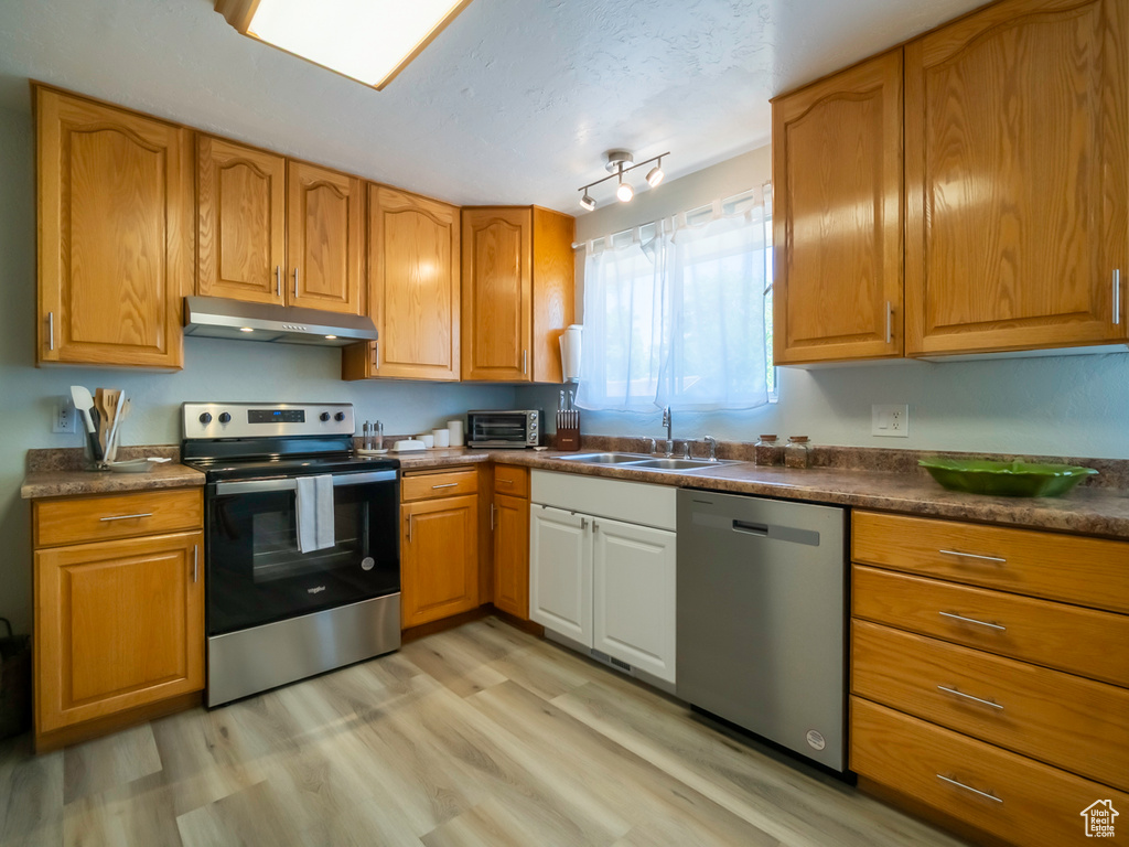 Kitchen featuring stainless steel appliances, under cabinet range hood, a sink, light wood-style floors, and dark countertops