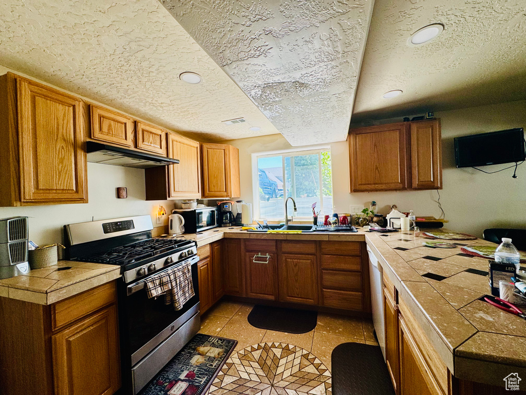 Kitchen featuring appliances with stainless steel finishes, a sink, under cabinet range hood, a textured ceiling, and brown cabinetry