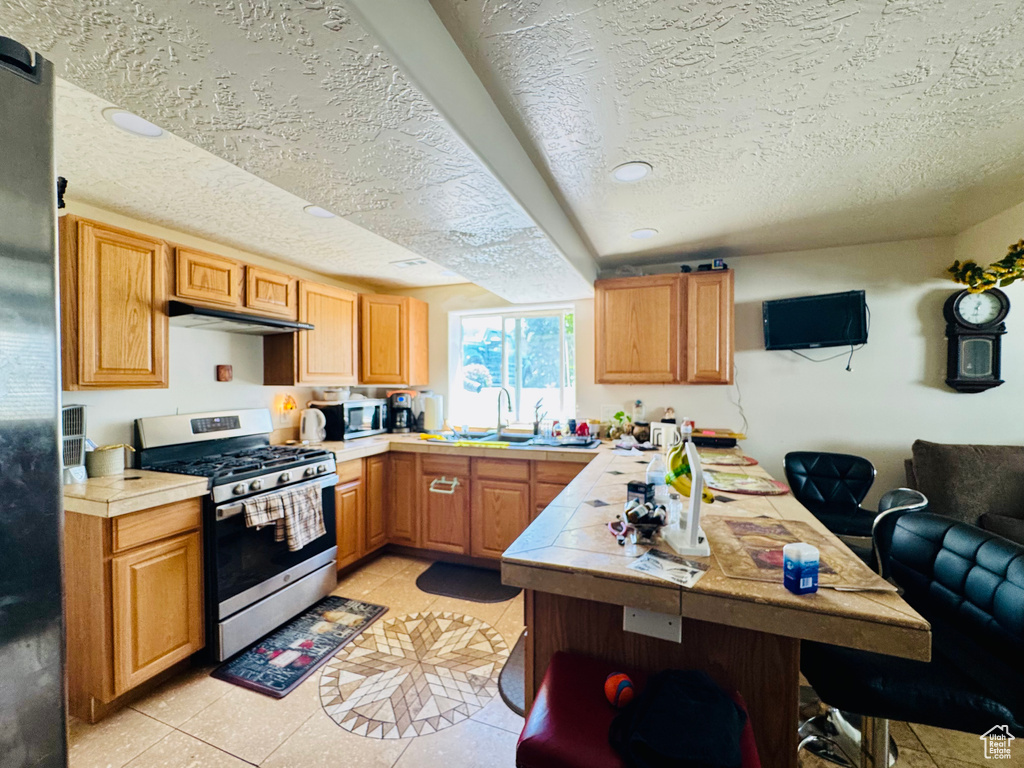 Kitchen with stainless steel appliances, a textured ceiling, tile countertops, under cabinet range hood, and a sink