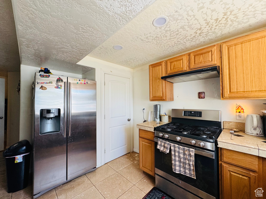 Kitchen with appliances with stainless steel finishes, under cabinet range hood, light countertops, light tile patterned floors, and a textured ceiling