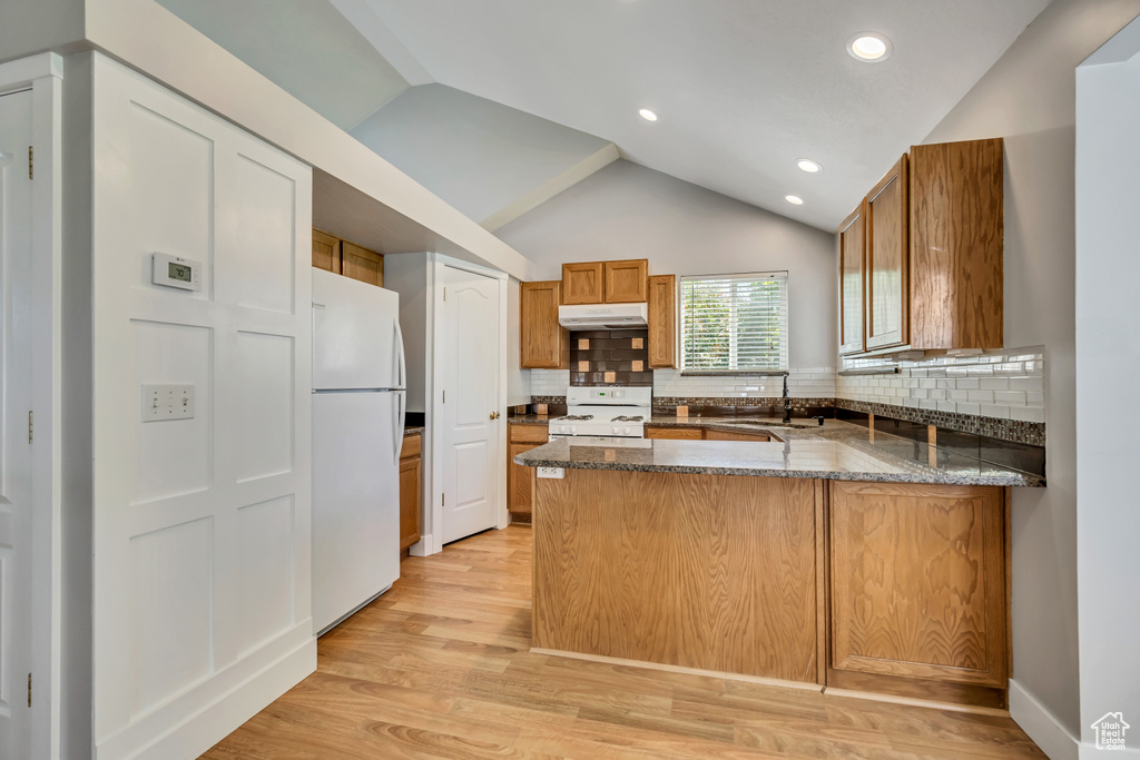 Kitchen featuring white appliances, under cabinet range hood, a peninsula, dark stone counters, and backsplash