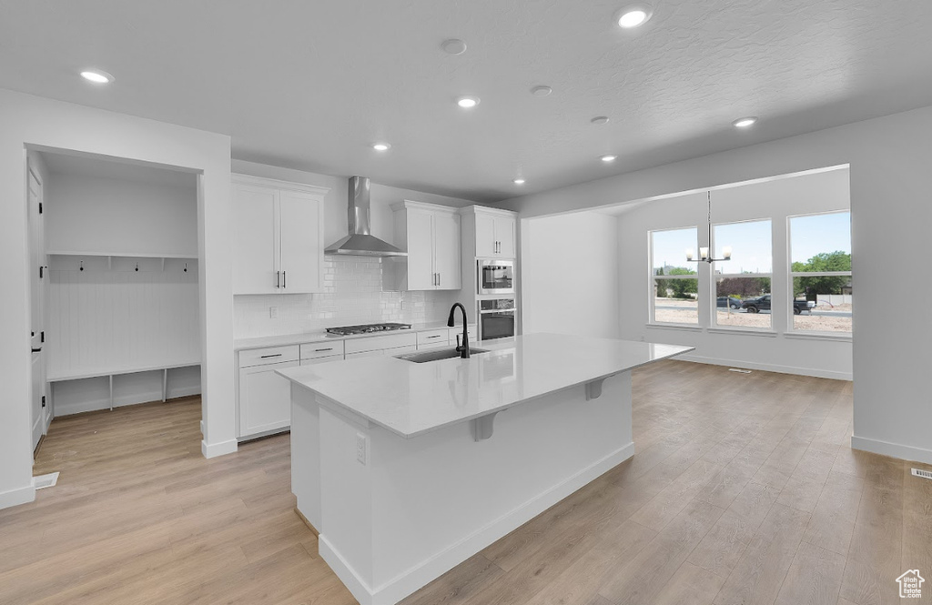 Kitchen featuring wall chimney exhaust hood, stainless steel appliances, an island with sink, light wood-style floors, and light countertops
