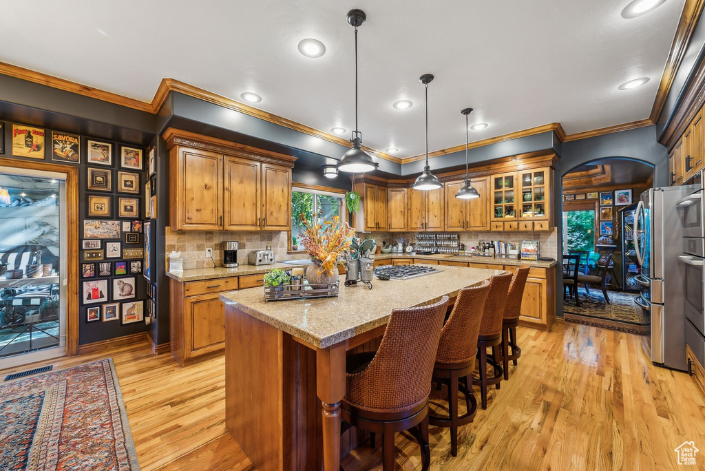 Kitchen featuring brown cabinets, ornamental molding, light wood finished floors, appliances with stainless steel finishes, and tasteful backsplash