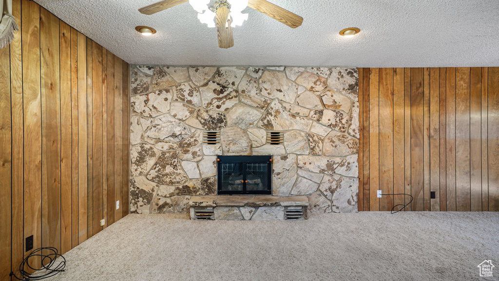 Unfurnished living room featuring wood walls, carpet flooring, a textured ceiling, a ceiling fan, and a stone fireplace