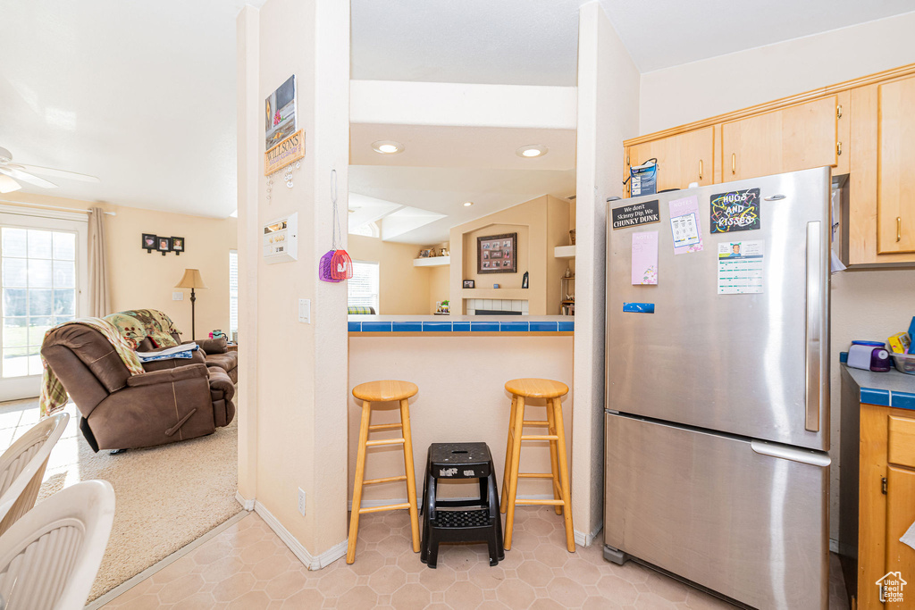 Kitchen featuring freestanding refrigerator, tile counters, light brown cabinetry, a ceiling fan, and a kitchen bar