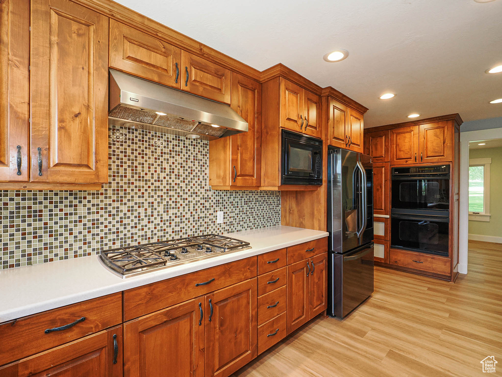 Kitchen featuring black appliances, under cabinet range hood, brown cabinetry, light countertops, and backsplash