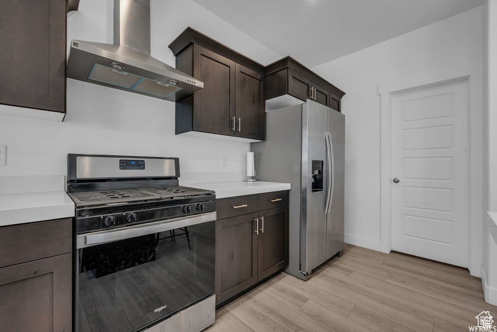 Kitchen featuring stainless steel appliances, wall chimney exhaust hood, light wood-style flooring, dark brown cabinetry, and light countertops