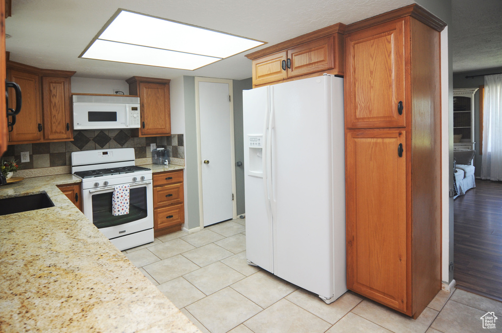 Kitchen with white appliances, tasteful backsplash, light tile patterned floors, brown cabinets, and light stone counters