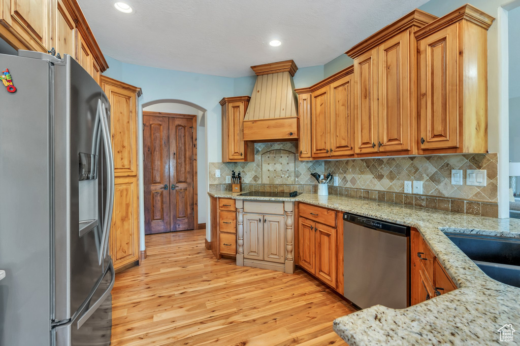 Kitchen featuring appliances with stainless steel finishes, light wood-style floors, custom range hood, light stone countertops, and decorative backsplash