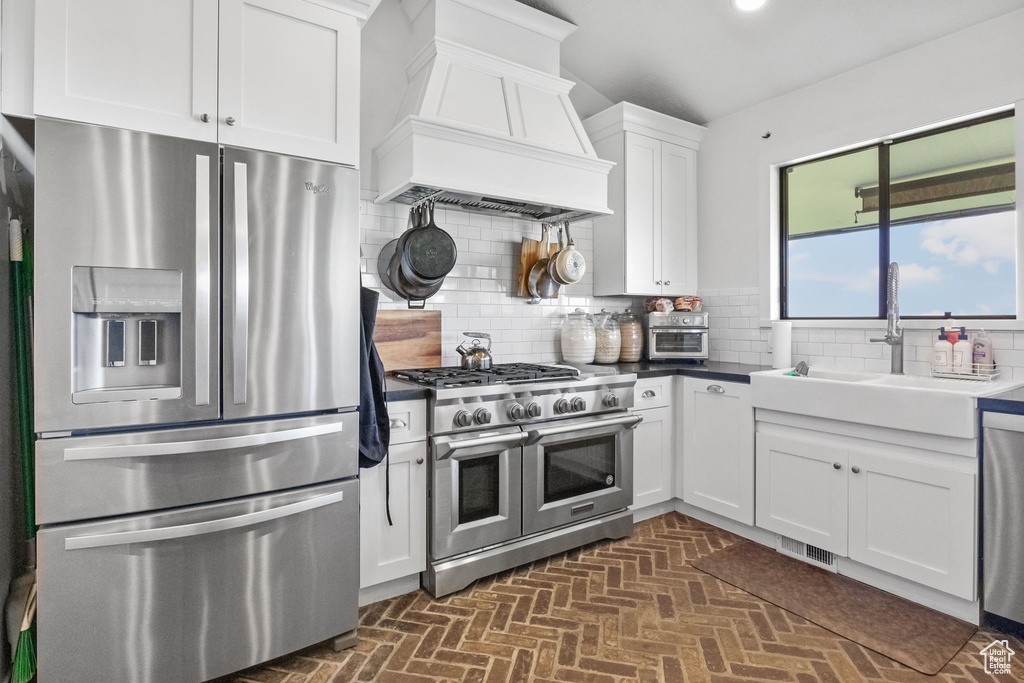 Kitchen with stainless steel appliances, custom exhaust hood, a sink, backsplash, and white cabinetry