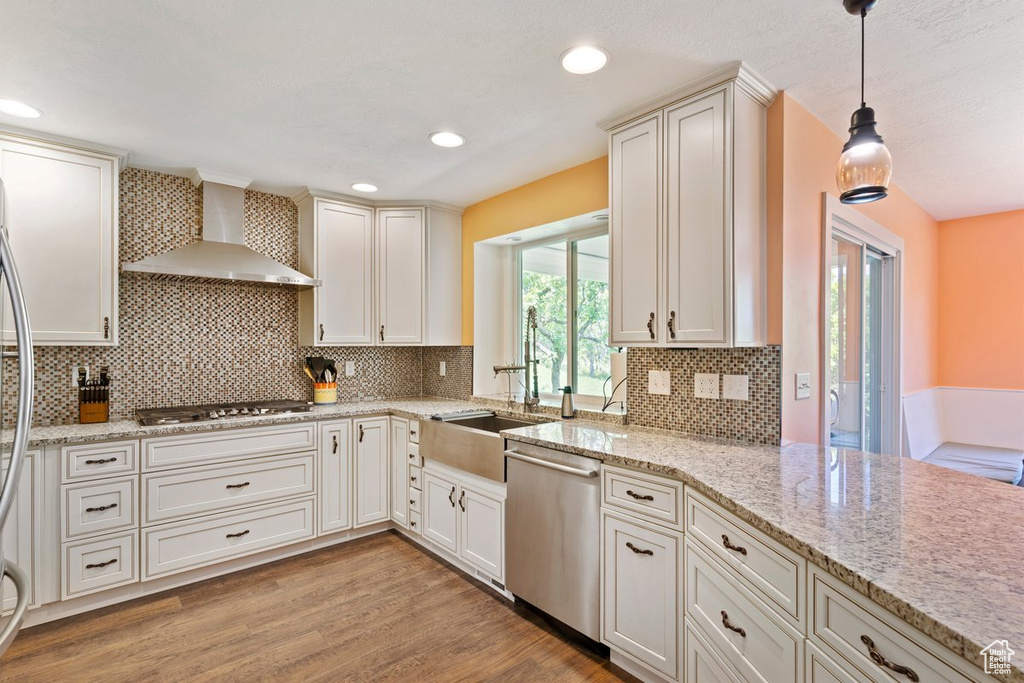 Kitchen with wall chimney range hood, appliances with stainless steel finishes, light wood-style flooring, hanging light fixtures, and light stone counters