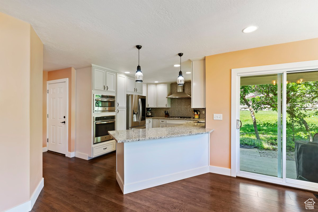 Kitchen with stainless steel appliances, wall chimney exhaust hood, a peninsula, light stone counters, and dark wood-type flooring