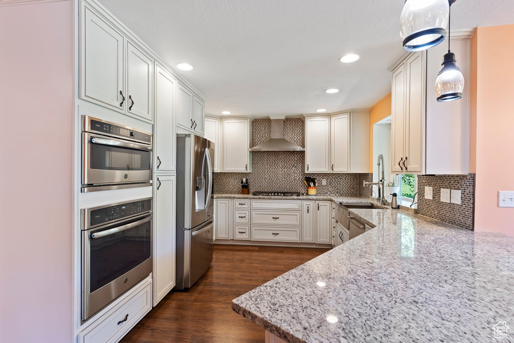 Kitchen with stainless steel appliances, wall chimney range hood, light stone counters, dark wood-style floors, and decorative backsplash