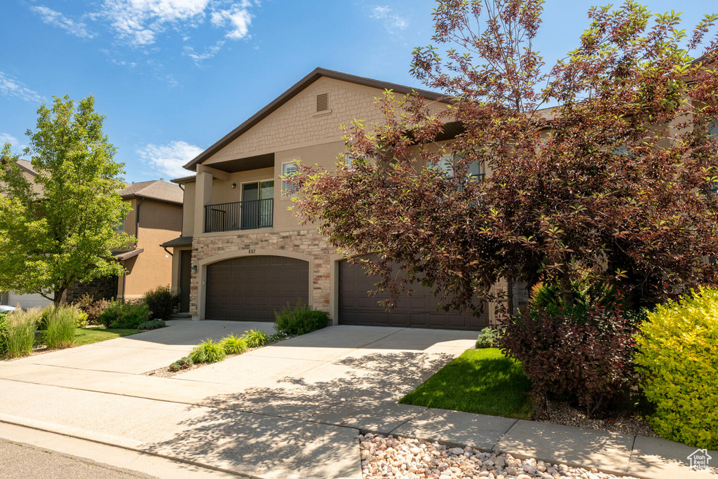 View of property with driveway, stucco siding, a balcony, a garage, and stone siding