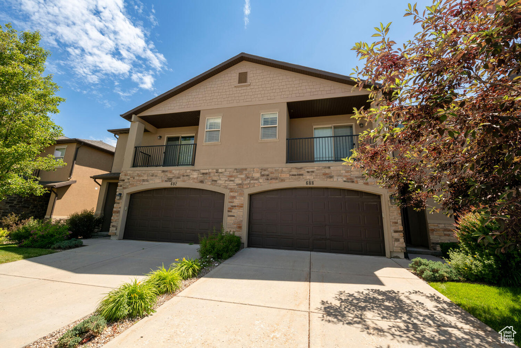 View of front facade featuring driveway, stucco siding, an attached garage, stone siding, and a balcony