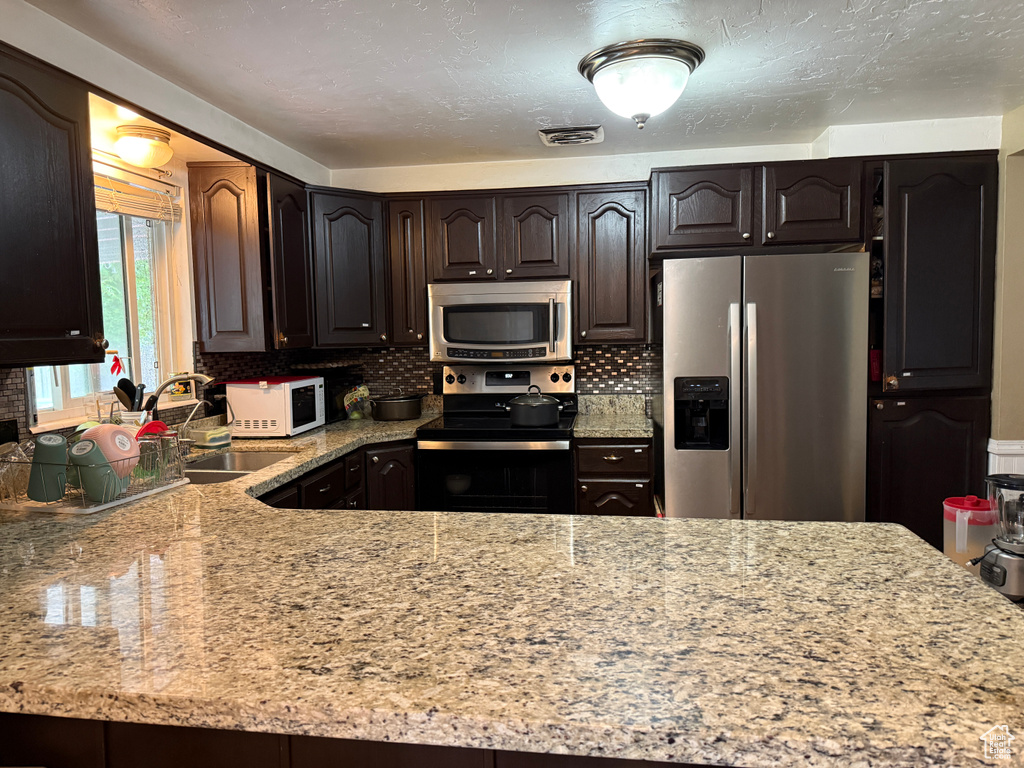 Kitchen with appliances with stainless steel finishes, light stone counters, a sink, dark brown cabinetry, and decorative backsplash