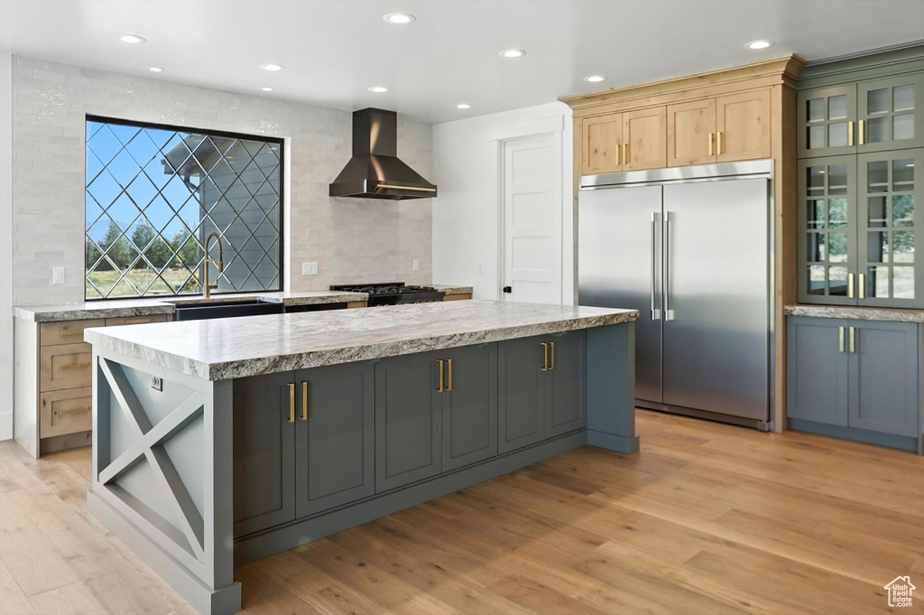 Kitchen featuring wall chimney exhaust hood, built in fridge, backsplash, light wood-style flooring, and a kitchen island