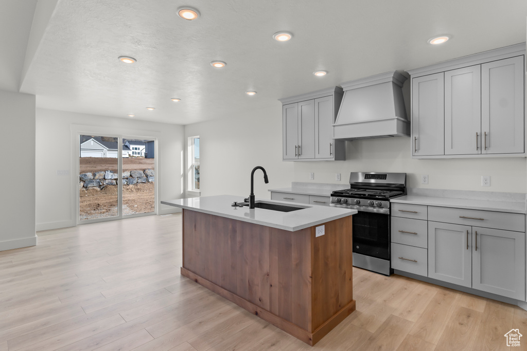 Kitchen featuring premium range hood, gas range, a sink, gray cabinets, and an island with sink