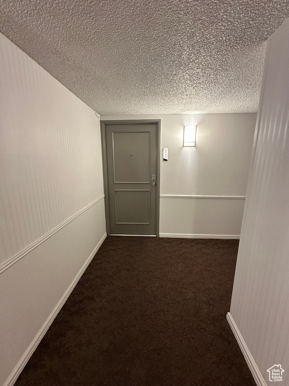 Hallway with a textured ceiling, dark colored carpet, and baseboards