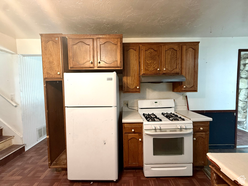 Kitchen with white appliances, under cabinet range hood, light countertops, and brown cabinets