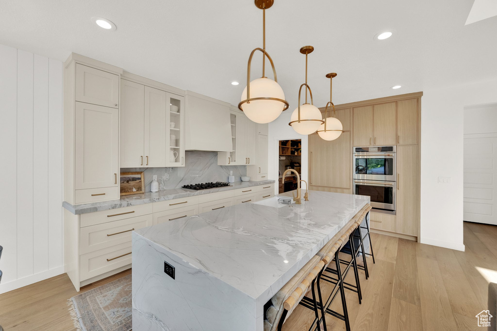 Kitchen featuring a sink, stainless steel double oven, light wood finished floors, an island with sink, and glass insert cabinets