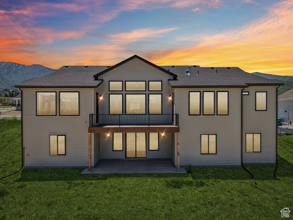 Back of property at dusk with a patio area, a balcony, a lawn, and a shingled roof