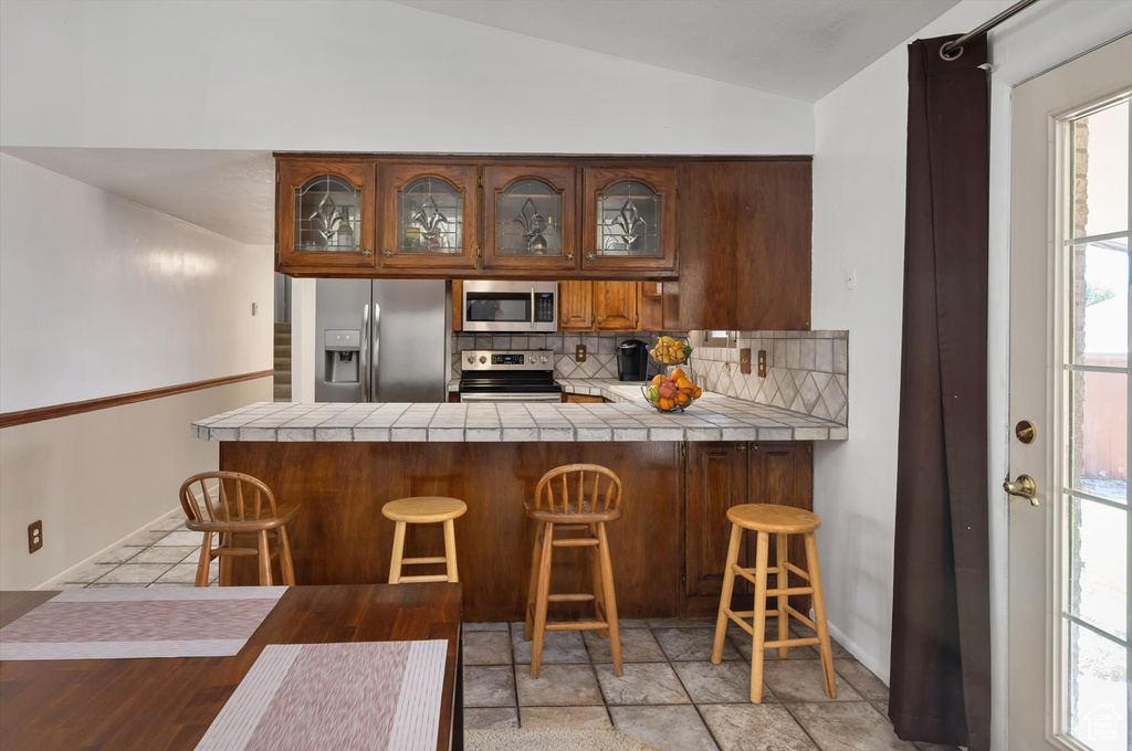 Kitchen featuring stainless steel appliances, backsplash, tile countertops, glass insert cabinets, and vaulted ceiling