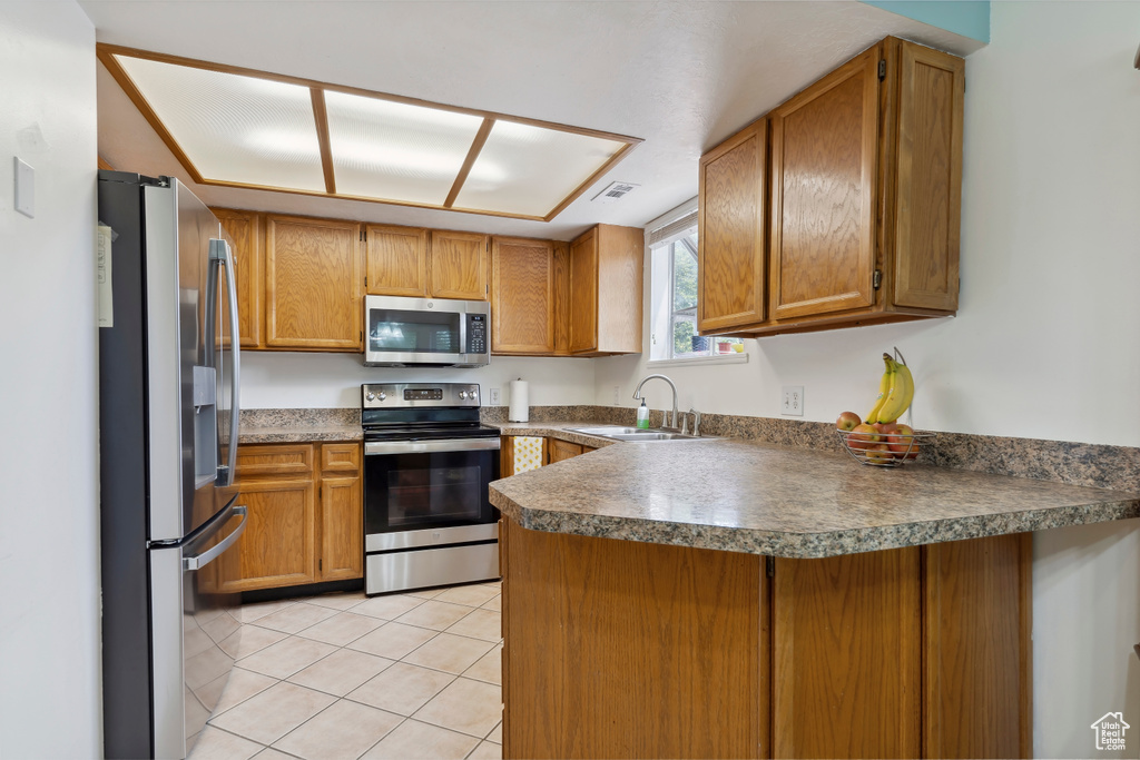 Kitchen with appliances with stainless steel finishes, a sink, a peninsula, light tile patterned floors, and brown cabinets