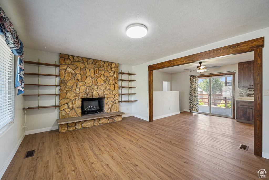 Unfurnished living room featuring wood finished floors, baseboards, a stone fireplace, a ceiling fan, and a textured ceiling
