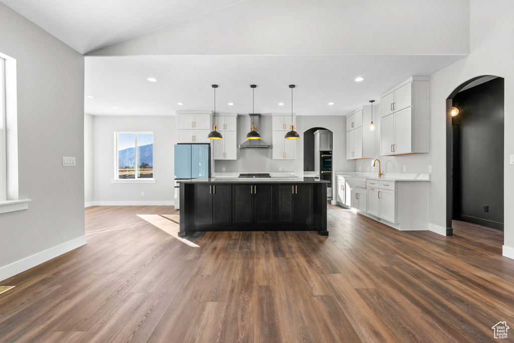 Kitchen featuring wall chimney exhaust hood, arched walkways, white cabinetry, recessed lighting, and vaulted ceiling