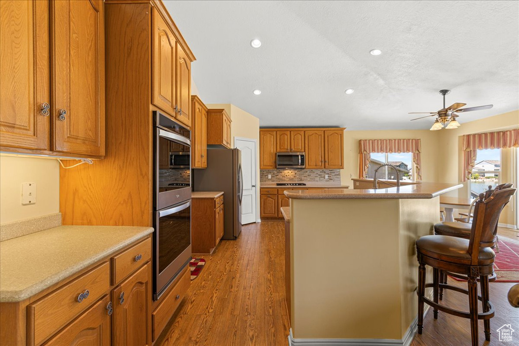 Kitchen featuring appliances with stainless steel finishes, a kitchen island with sink, brown cabinets, recessed lighting, and tasteful backsplash