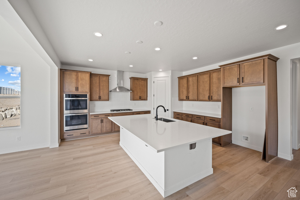 Kitchen with wall chimney exhaust hood, stainless steel double oven, brown cabinets, a sink, and recessed lighting