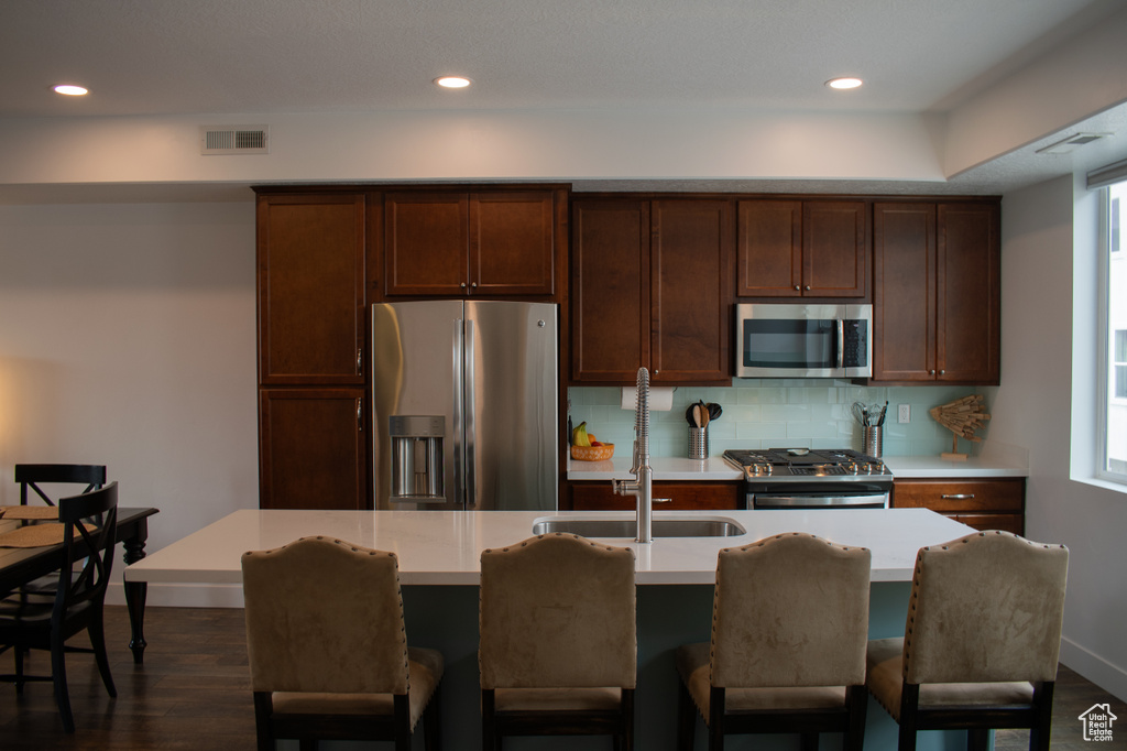 Kitchen with appliances with stainless steel finishes, light countertops, dark wood-type flooring, backsplash, and a center island with sink