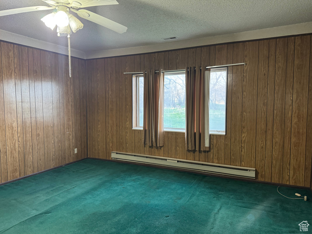 Carpeted empty room with a baseboard heating unit, wooden walls, ceiling fan, and a textured ceiling