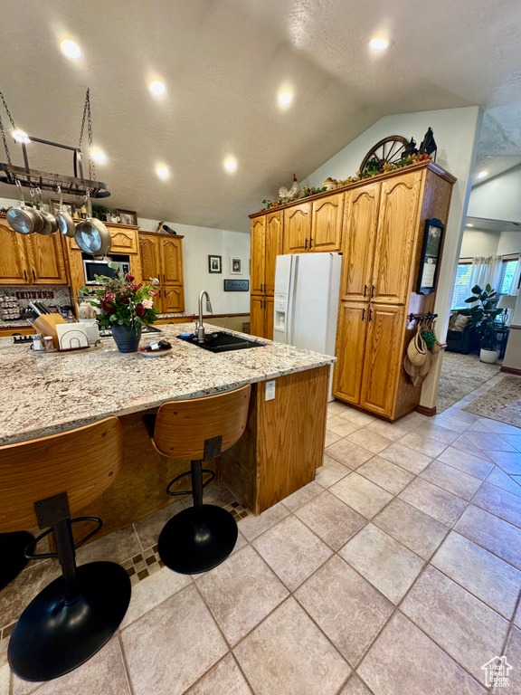 Kitchen with white refrigerator with ice dispenser, a sink, vaulted ceiling, light stone counters, and brown cabinetry