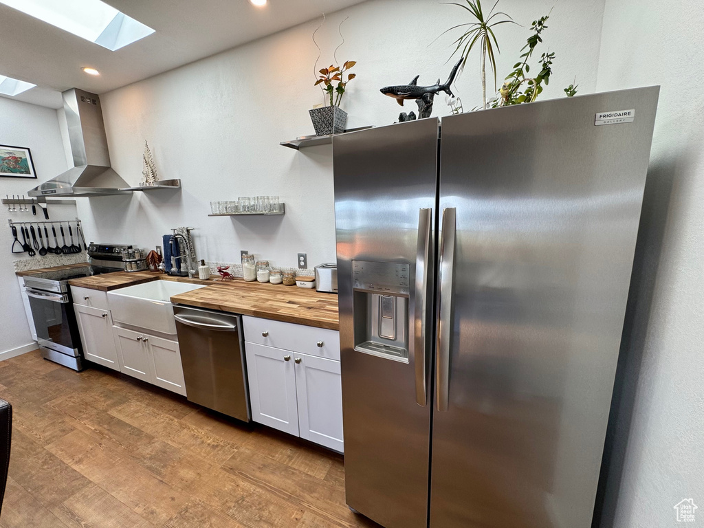 Kitchen featuring stainless steel appliances, butcher block countertops, open shelves, a skylight, and wall chimney exhaust hood