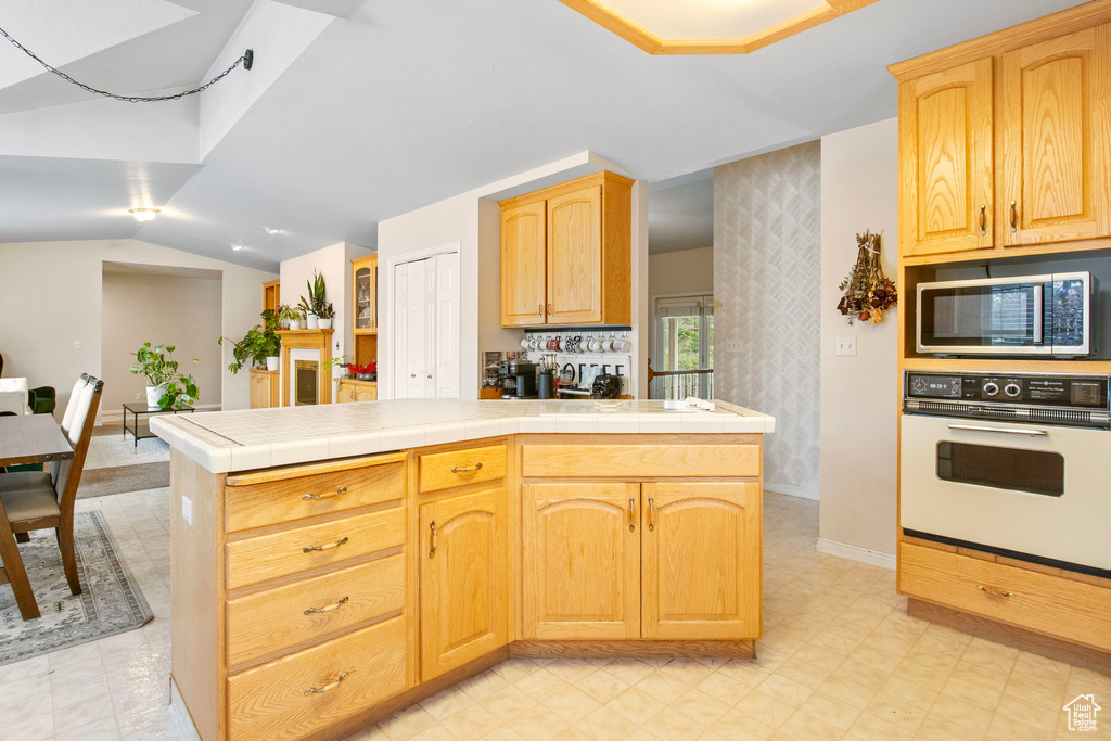 Kitchen featuring white oven, stainless steel microwave, vaulted ceiling, tile countertops, and light brown cabinetry