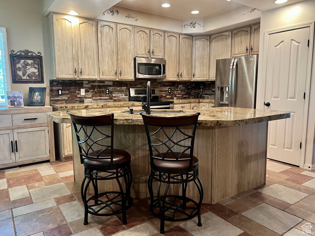 Kitchen with appliances with stainless steel finishes, tasteful backsplash, a center island with sink, a breakfast bar, and light stone finish flooring