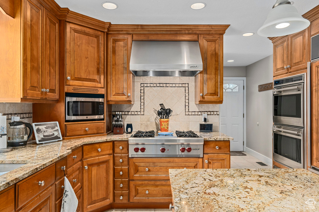 Kitchen with wall chimney exhaust hood, appliances with stainless steel finishes, backsplash, brown cabinetry, and recessed lighting