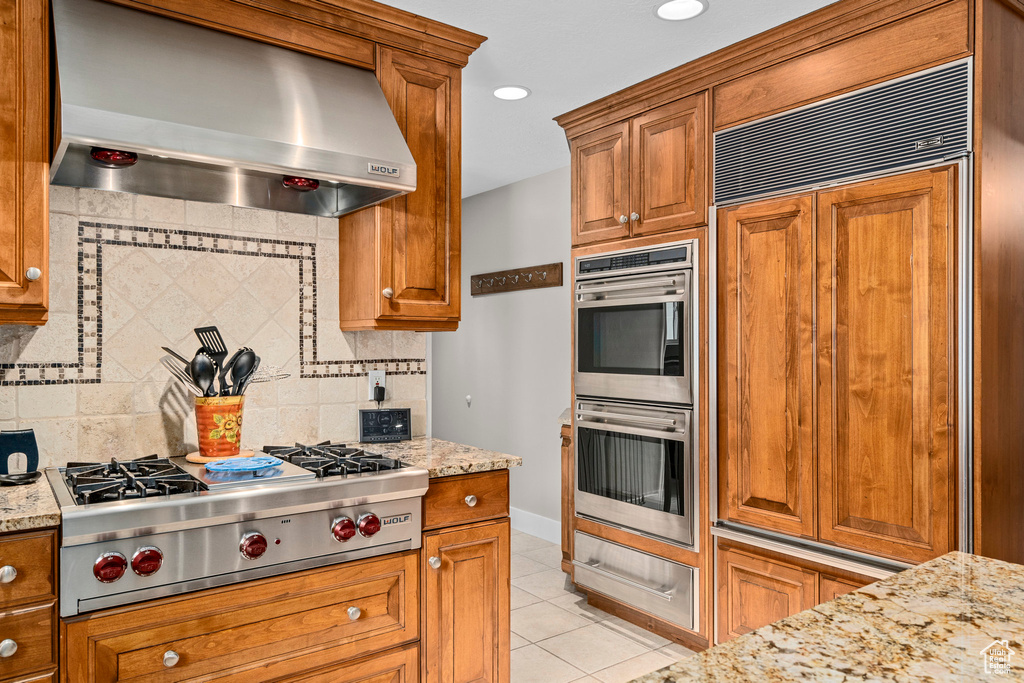 Kitchen featuring wall chimney range hood, stainless steel appliances, backsplash, light tile patterned floors, and brown cabinetry
