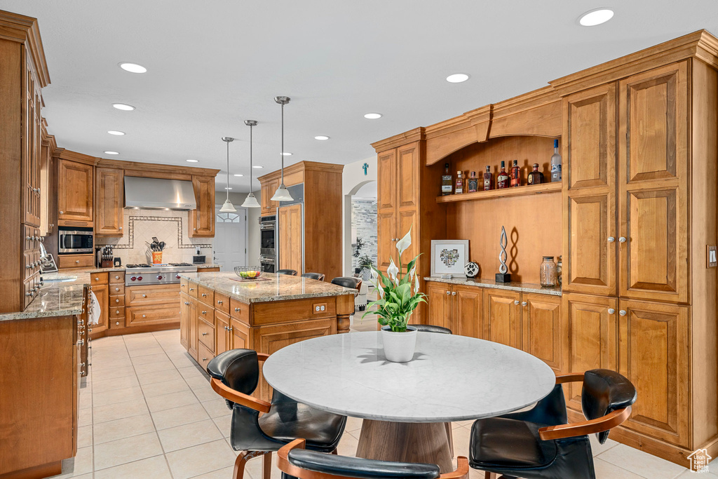 Kitchen featuring wall chimney exhaust hood, appliances with stainless steel finishes, a kitchen island, brown cabinets, and light tile patterned floors