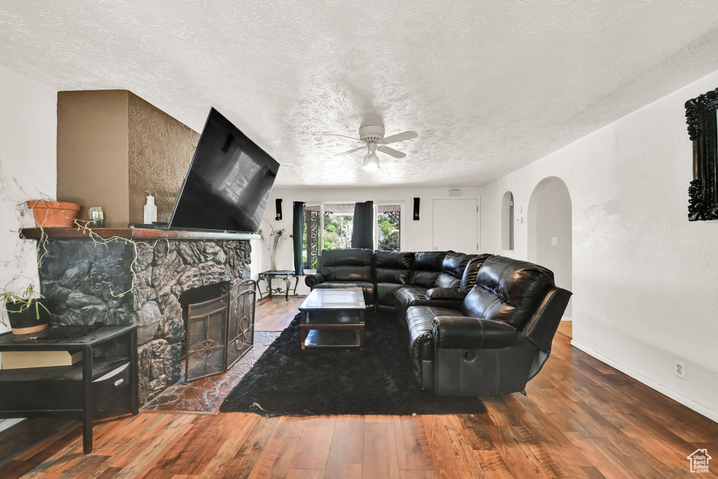 Living area with arched walkways, ceiling fan, wood-type flooring, a textured ceiling, and a stone fireplace