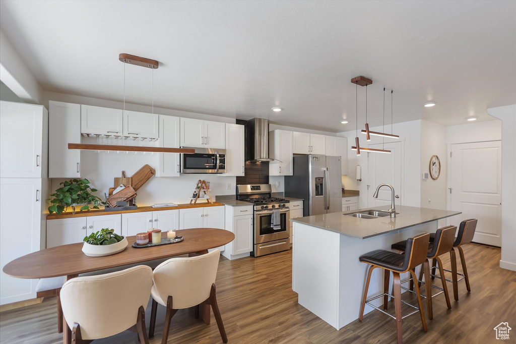 Kitchen with stainless steel appliances, a sink, wall chimney exhaust hood, a kitchen island with sink, and wood finished floors