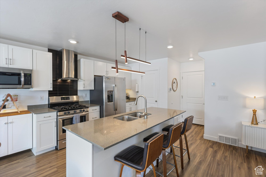 Kitchen featuring appliances with stainless steel finishes, a sink, wall chimney exhaust hood, dark wood-style flooring, and a kitchen bar