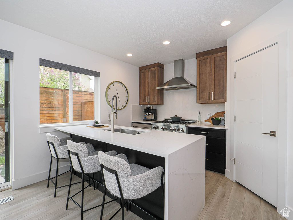 Kitchen with wall chimney exhaust hood, range, light wood-style floors, a center island with sink, and recessed lighting