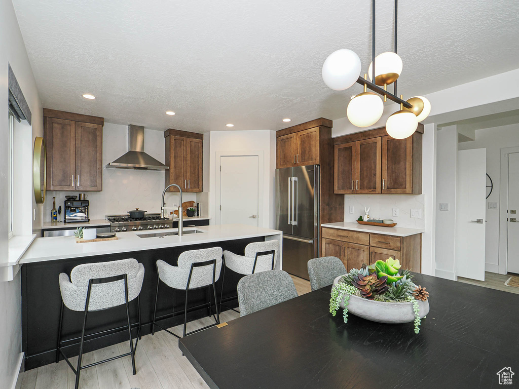 Kitchen with high end fridge, wall chimney exhaust hood, a sink, a breakfast bar, and light wood finished floors